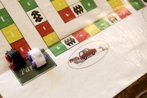 Foto de parte de um jogo de tabuleiro. No local escrito "partida", há dois carrinhos de plástico.