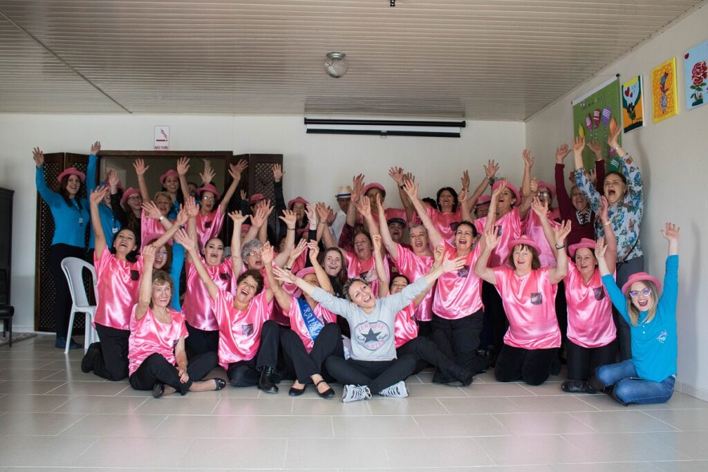 Foto de um grupo de pessoas com roupas, em sua maioria, na cor rosa, com as mãos para cima. Elas estão sorrindo.