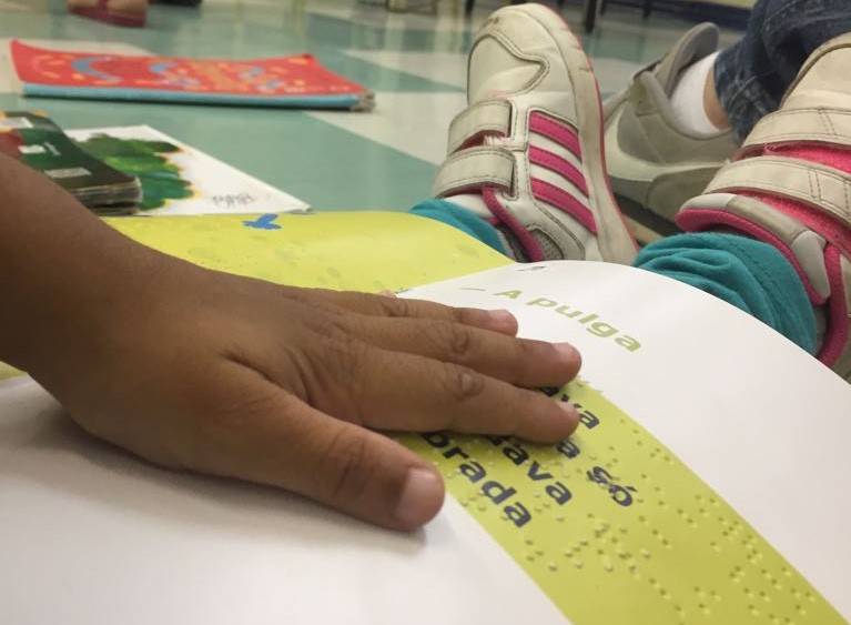 Foto em close da mão de uma criança lendo um livro em braile sentada no chão
