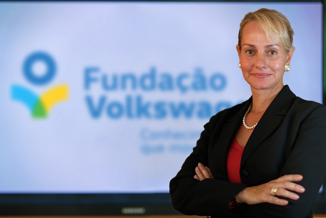 Foto de uma mulher branca, de aproximadamente 40 anos, com cabelos loiros presos em um coque e braços cruzados. Em uma sala fechada, com um telão ao fundo com o logo da Fundação Volkswagen