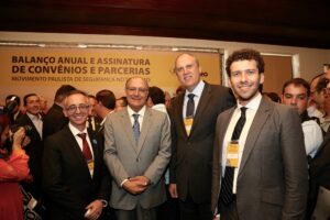 Foto de quatro homens pé em um ambiente fechado. Eles sorriem e estão usando terno e gravata.