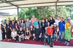 Mais de trinta pessoas reunidas em um ambiente externo com cobertura. Elas sorriem. Algumas estão em pé e outras estão agachadas.