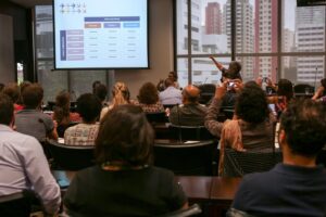 Foto de duas mulheres na frente de uma plateia lotada. Uma delas aponta para um telão com uma tabela.