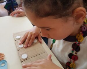 Foto de uma menina sentada colocando as duas mãos em pequenos círculos brancos em alto relevo. Eles estão em cima de pedaços de papelão em uma mesa.