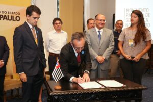 Foto de um senhor assinando um papel em cima de uma mesa em uma sala com várias pessoas em pé.