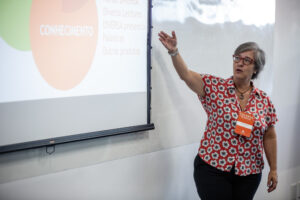Foto de uma mulher em pé, mostrando um telão com a mão direita em uma sala.