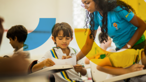 Foto de uma professora e algumas crianças em sala de aula. A professora está em pé e ajuda um dos alunos.