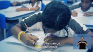 Foto de uma garota na sala de aula. Ela está debruçada sobre o caderno e escreve nele.