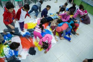 Crianças brincam sobre um grande filme plástico estendido no chão da sala de aula. Ele tem, na parte interior, diversas manchas de tinta coloridas