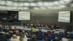 Visão do auditório com o público sentado em fileiras e, ao fundo, palco com mesa e debatedores.