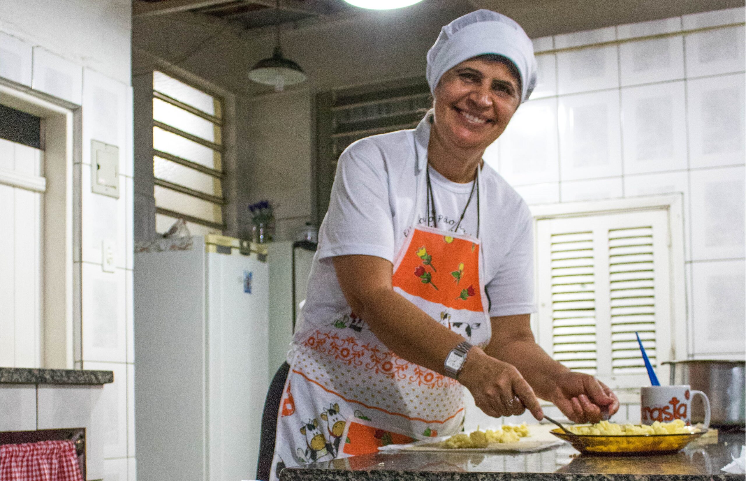 Senhora sorri enquanto manipula alimentos na cozinha da organização social Comunidade Brilho Sagrada Família