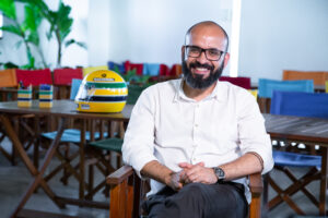 Helton Souto Lima, homem negro careca, com baba cheia e óculos com armação preta. Ele está sentado e sorri