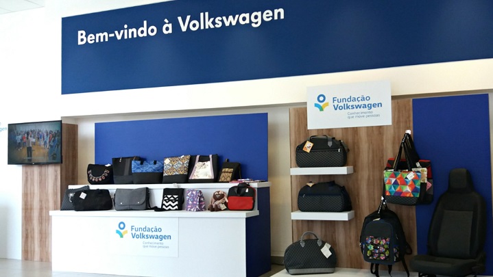 Foto em plano aberto, com um balcão e prateleiras diversas com bolsas e malas expostos. No topo, em azul, há o texto: "Bem-vindo à Volkswagen". No gabinete do balcão, há o logotipo da Fundação Volkswagen.