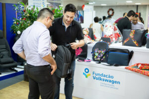 Visão geral de visitantes no Bazar Social da Fundação Volkswagen. À frente, dois homens manipulam mochila. Ao fundo, estante com produtos diversos, espelho e árvore de Natal.