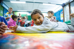 Sob tapete colorido com tinta, crianças brincam. Destaque para um menino deitado de bruços, que sorri.