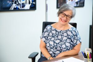 Liliane Garcez está sentada junto a uma mesa e sorri para a câmera, com a cabeça de lado.