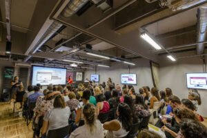 Visão de fundo de um auditório que está com as cadeiras lotadas de pessoas.