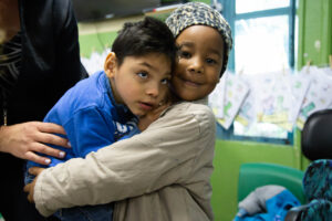 Menina beneficiada pelo projeto Brincar sorri para a câmera enquanto abraça colega de classe com paralisia cerebral.