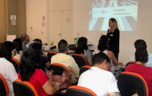 Auditório com poltronas laranjas dispostas lado a lado, de costas para a câmera. Ao fundo, à frente da sala, instrutora em pé junto a uma projeção com o título Cidadania em Movimento em destaque.