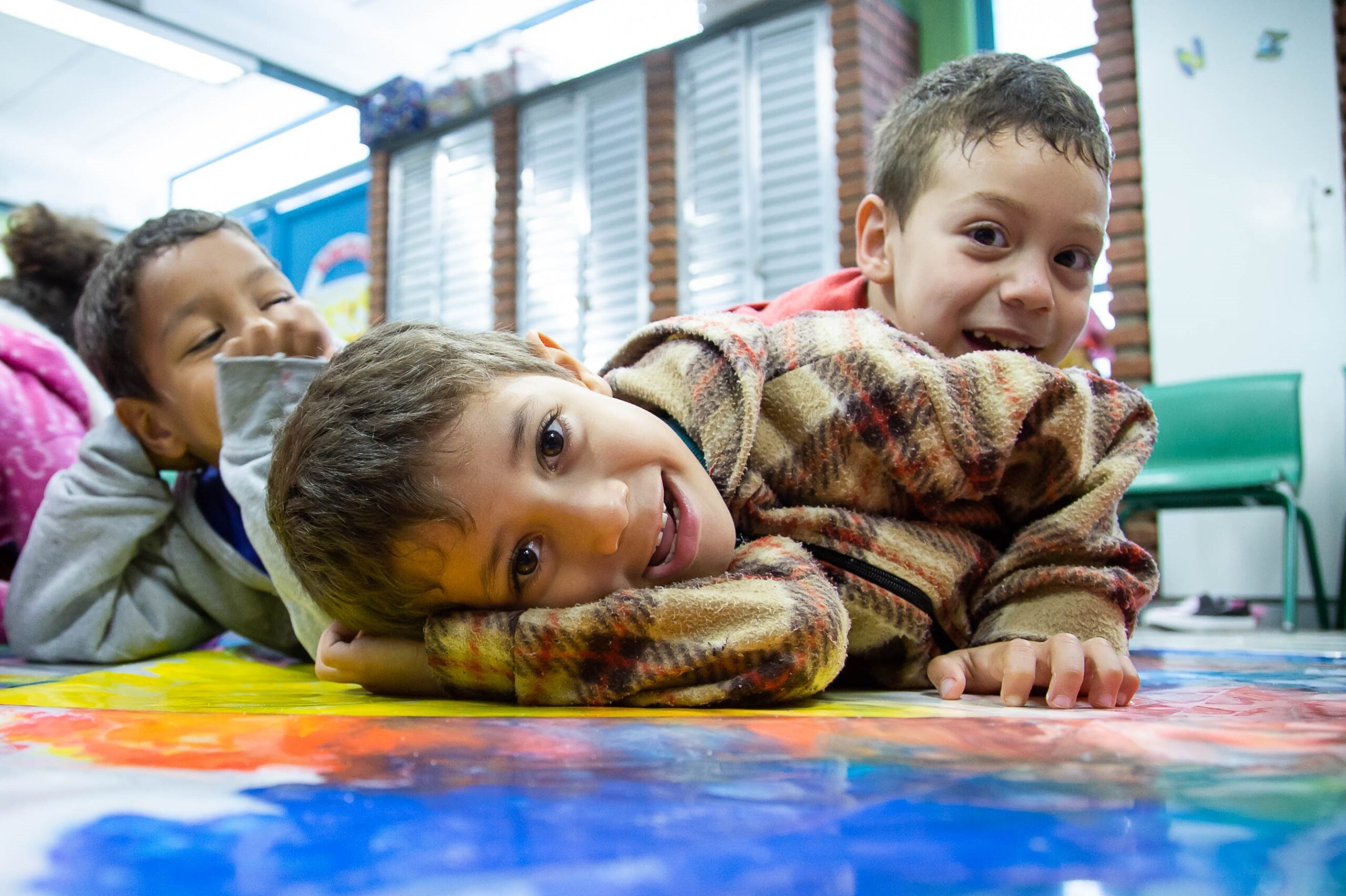 Crianças sorriem para câmera deitadas sobre um tapete colorido.