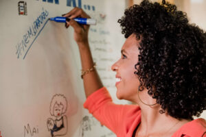 Mulher negra sorri enquanto escreve sobre um quadro branco a hashtag #Nem1PraTrás