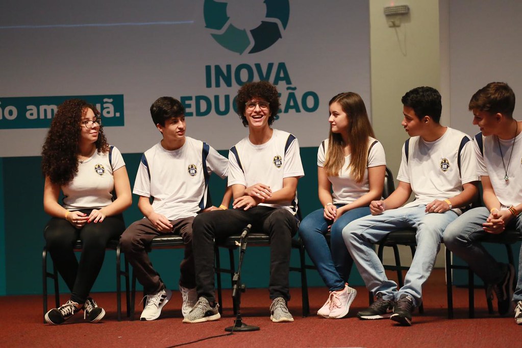 Adolescentes sentados em cadeiras num palco, com painel ao fundo escrito "Inova Educação"