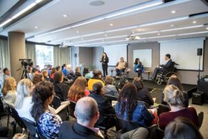 Visão de auditório com homens e mulheres sentados em fileiras, de costas para a câmera. Ao fundo, dois telões e palco, com jornalista sentada ao centro, dois cadeirantes ao lado e intérprete de LIBRAS em pé à esquerda da imagem.