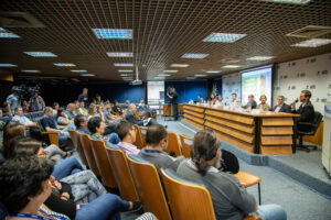 Visão de auditório com poltronas de madeira e mesa com autoridades e telão à frente.