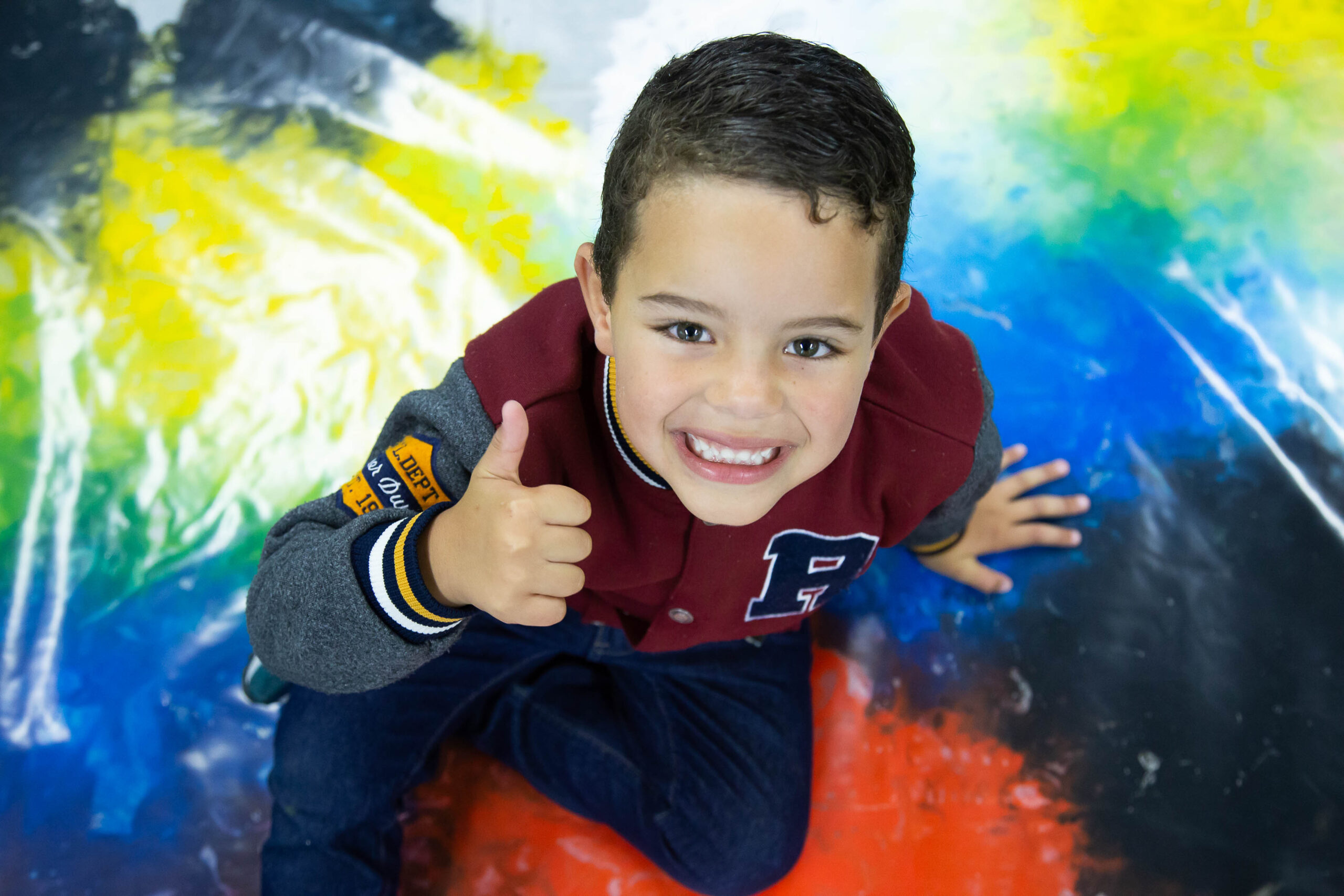 Foto de um menino visto de cima. Ele está sorrindo e faz o sinal de positivo com a mão direita.