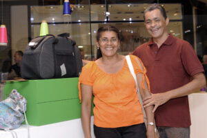 Casal sorri para a câmera em meio a vitrines com bolsa e outros produtos.