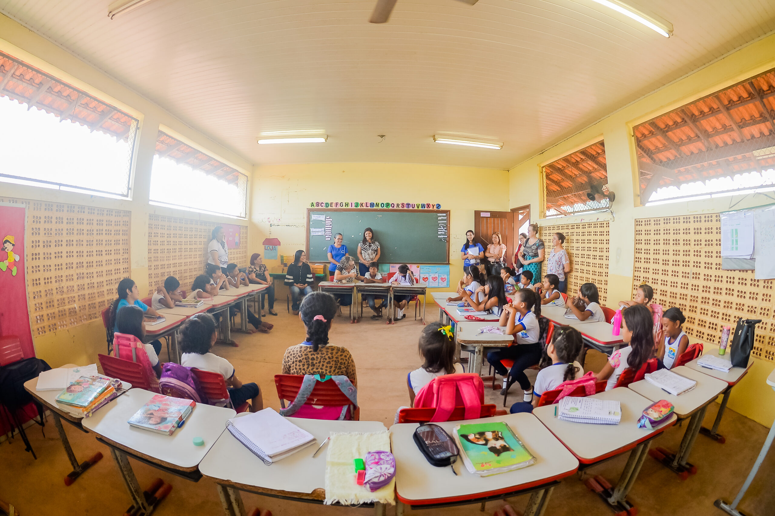 Foto de um grupo diverso de crianças sentadas em uma sala de aula. Há um grupo de adultos em pé próximos a lousa.