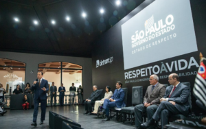 Auditório com pessoas sentadas. Ao fundo, homem em pé discursando à esquerda e tela com o texto Respeito à vida