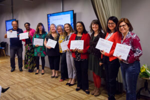 Foto de um grupo de pessoas em pé, sendo um homem e oito mulheres, em uma sala. Eles estão parados um do lado do outro e seguram um certificado nas mãos. Todos estão sorrindo.