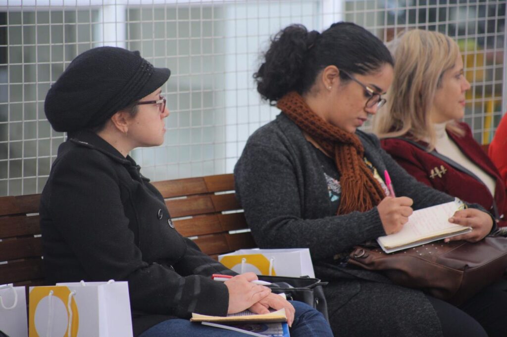 Foto de uma mulher fazendo anotação em um caderno. Ao seu lado, há duas mulheres.