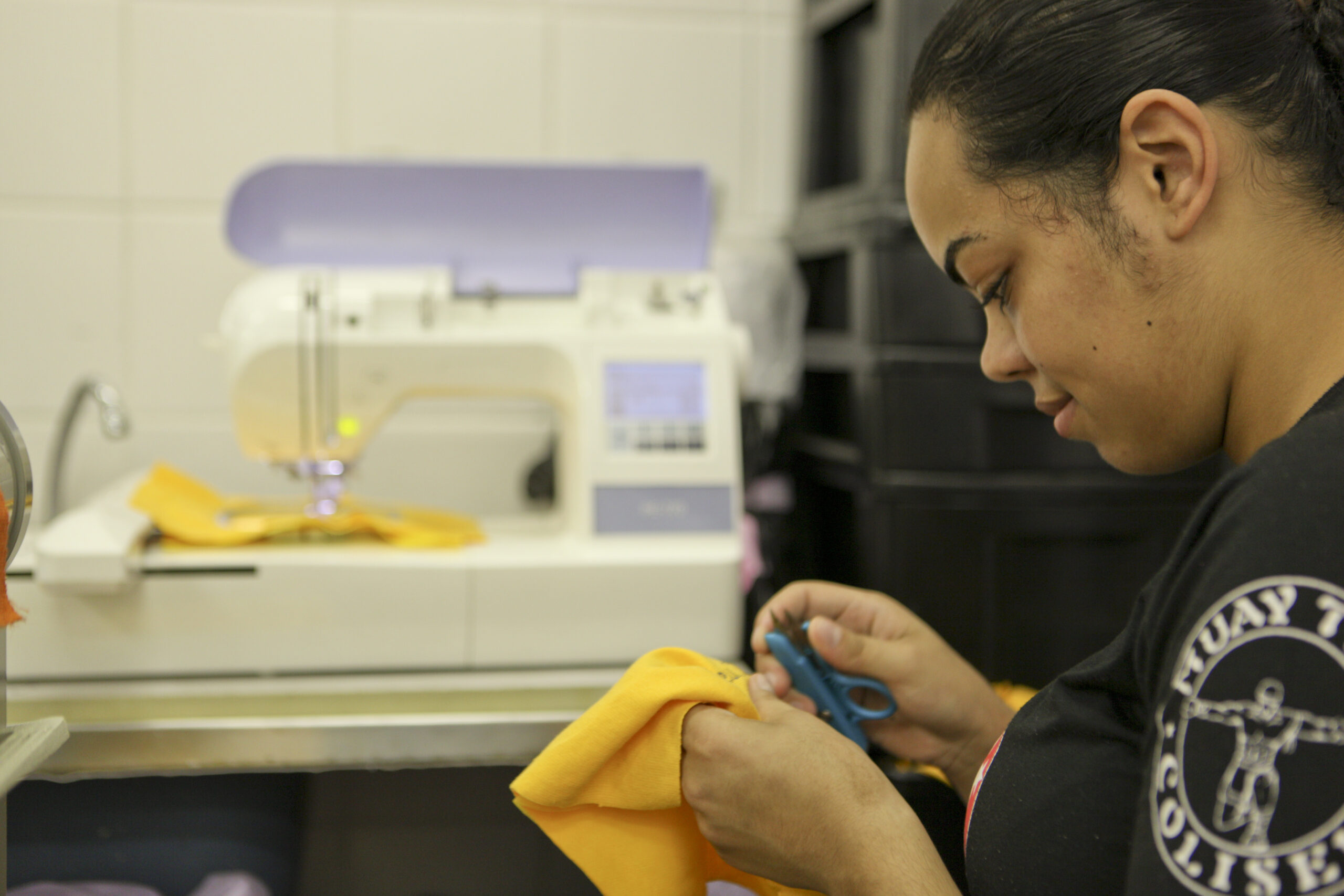 Foto de uma mulher jovem segurando uma tesoura e um tecido nas mãos. Ao lado dela, há uma máquina de costura.