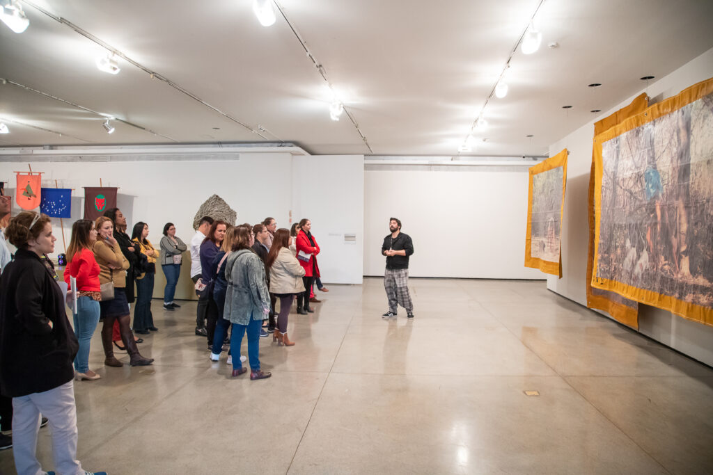 Foto de um grupo de pessoas em pé em uma galeria de artes. Um homem fala para o grupo