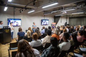Em auditório, educadores sentados, de costas, assistem à aula do projeto.