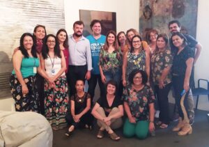 Foto de um grupo diverso de mulheres e homens, sendo que alguns estão em pé e outros sentados no chão, sorriem para a foto