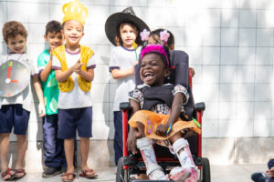 Foto de um grupo diverso de crianças fantasiadas em pé em um pátio. Uma delas, está em uma cadeira de rodas.