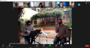 Print de uma vídeo chamada. No centro, há um homem sentado cantando enquanto outro toca teclado