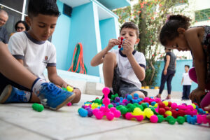 Foto de um grupo de crianças sentados no pátio de uma escola. Eles estão brincando com objetos coloridos.